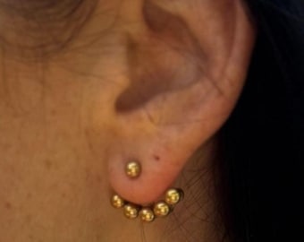 May include: Gold-colored ear jacket earrings. The earrings feature a small stud in the earlobe and a curved row of six small, round beads that sit behind the ear. The jewelry is shown on a person's ear.