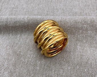 May include: A gold-colored ring with a thick, layered design. The ring features multiple bands of metal, creating a textured and dimensional appearance. The ring is set against a neutral gray background.