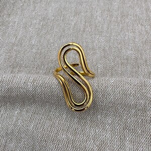 Könnte beinhalten: Ein goldfarbener Ring mit einem einzigartigen, abstrakten Design. Der Ring hat ein fließendes, offenes Muster mit geschwungenen Linien und einer polierten Oberfläche. Der Ring ist vor einem neutralgrauen Stoffhintergrund platziert.