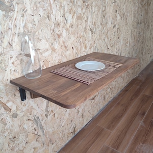 Peut inclure: Une table murale rectangulaire en bois aux coins arrondis, dot&eacute;e d'une finition en grain de bois brun fonc&eacute;. Une carafe en verre transparent est pos&eacute;e &agrave; gauche, tandis qu'une assiette en c&eacute;ramique blanche repose sur un set de table en bambou.