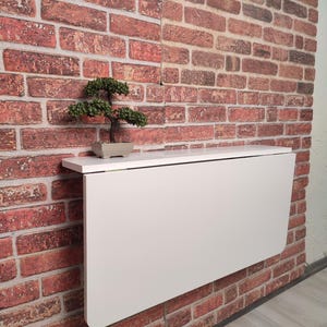 Könnte beinhalten: Ein weißer, klappbarer Wandtisch mit abgerundeter Kante. Der Tisch ist an einer Backsteinwand montiert und hat ein kleines, eingetopftes Bonsai-Bäumchen auf dem Regal darüber.
