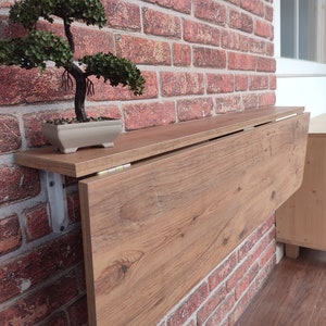 May include: A brown wooden folding table mounted to a brick wall. The table is folded up and has a light wood grain finish. A small potted bonsai tree is on the shelf above the table.