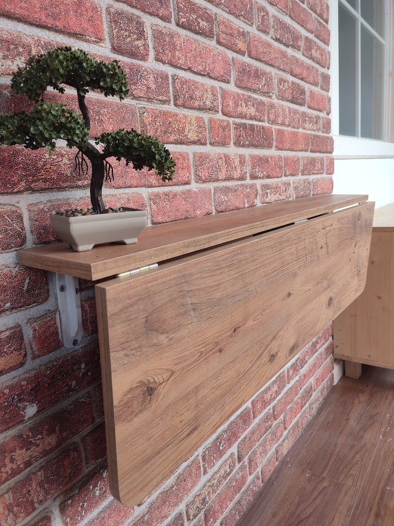 Może przedstawiać: Brązowy drewniany st&oacute;ł składany zamontowany na ścianie z cegły. St&oacute;ł jest złożony i ma jasne usłojenie drewna. Małe bonsai w doniczce znajduje się na p&oacute;łce nad stołem.