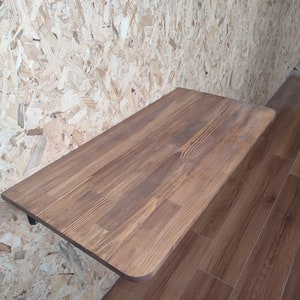 Peut inclure: Un bureau rectangulaire fix&eacute; au mur avec une surface en bois teint&eacute; brun moyen, pr&eacute;sentant des motifs de grain visibles et des coins arrondis. Le bureau est fix&eacute; au mur par un support en m&eacute;tal noir, offrant un espace de travail plat et solide.