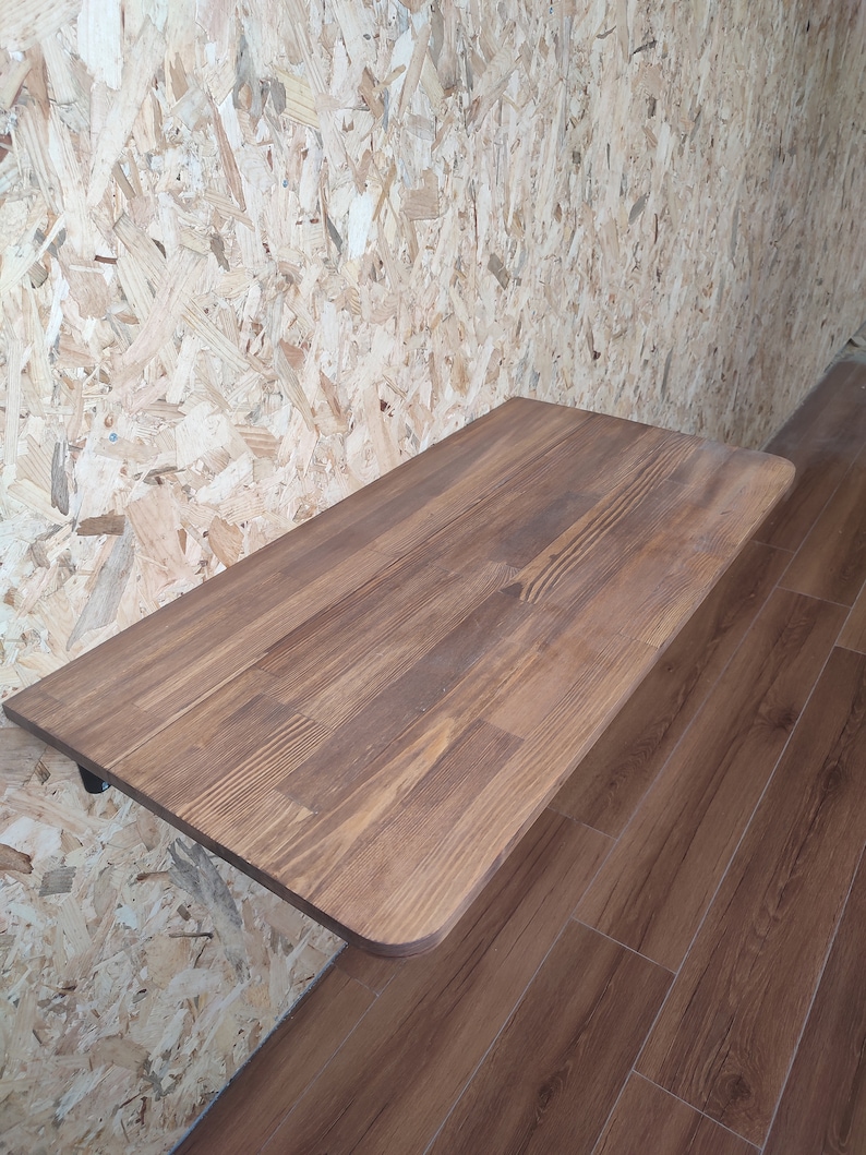 Peut inclure: Un bureau rectangulaire fix&eacute; au mur avec une surface en bois teint&eacute; brun moyen, pr&eacute;sentant des motifs de grain visibles et des coins arrondis. Le bureau est fix&eacute; au mur par un support en m&eacute;tal noir, offrant un espace de travail plat et solide.