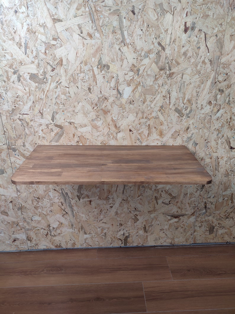 Peut inclure: Un bureau flottant rectangulaire fix&eacute; au mur, dot&eacute; d'une surface en bois brun fonc&eacute; au grain lisse et aux coins arrondis. Le bureau est fix&eacute; sur un mur recouvert de panneaux de particules orient&eacute;es clairs, cr&eacute;ant un contraste de tons.
