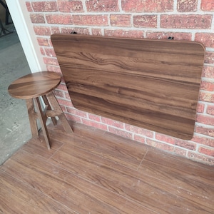 Puede incluir: Una mesa plegable de madera marrón montada en una pared de ladrillo. La mesa está cerrada y tiene un patrón de veta de madera. Un pequeño taburete redondo de madera está en primer plano.