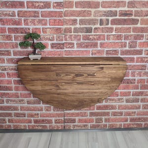 May include: A wall-mounted, half-circle wooden table with a dark brown finish. The table is mounted on a red brick wall. A small bonsai tree in a white pot sits on the table's surface.