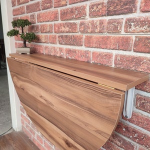 May include: A brown wooden folding wall table with a rounded edge, mounted to a brick wall. The table is folded up and has a small potted bonsai tree on the shelf above it.