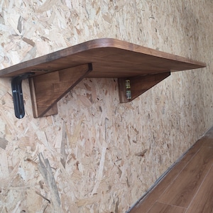 Peut inclure: Une table pliante murale dot&eacute;e d'une surface en bois rectangulaire aux coins arrondis. La table est soutenue par deux supports en bois et un m&eacute;canisme de charni&egrave;re en m&eacute;tal noir, permettant de replier la surface plane contre le mur.