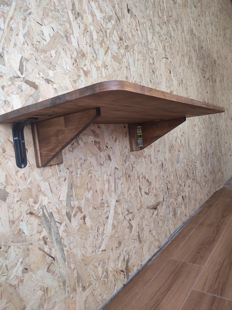 Peut inclure: Une table pliante murale dot&eacute;e d'une surface en bois rectangulaire aux coins arrondis. La table est soutenue par deux supports en bois et un m&eacute;canisme de charni&egrave;re en m&eacute;tal noir, permettant de replier la surface plane contre le mur.