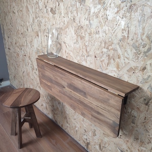 Peut inclure: Un bureau pliant mural en bois brun fonc&eacute; avec un tabouret rond assorti. Le bureau dispose d'un plateau articul&eacute; qui se replie vers le bas, et un vase en verre transparent est pos&eacute; sur la surface plane. Le mobilier est en bois vein&eacute;.