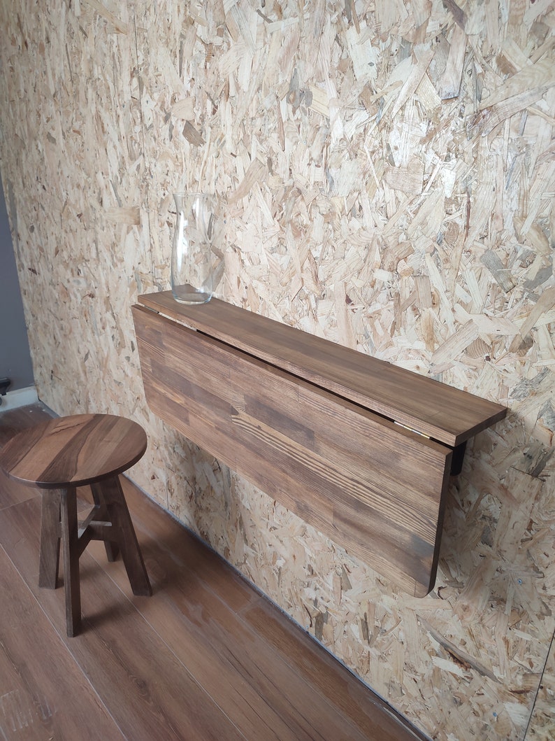 Peut inclure: Un bureau pliant mural en bois brun fonc&eacute; avec un tabouret rond assorti. Le bureau dispose d'un plateau articul&eacute; qui se replie vers le bas, et un vase en verre transparent est pos&eacute; sur la surface plane. Le mobilier est en bois vein&eacute;.