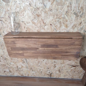Peut inclure: Une table pliante murale rectangulaire en bois brun avec un motif de grain de bois visible et une finition lisse. Une carafe en verre transparent est pos&eacute;e sur le c&ocirc;t&eacute; gauche de la surface sup&eacute;rieure. La table est repli&eacute;e contre le mur.