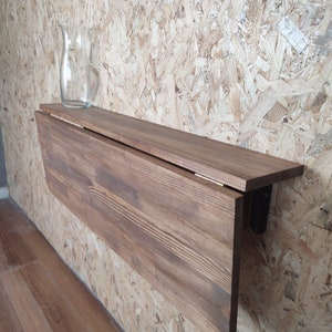 Peut inclure: Une table pliante murale en bois brun fonc&eacute; avec un plateau rectangulaire et un panneau rabattable articul&eacute;. Une carafe en verre transparent est pos&eacute;e sur le c&ocirc;t&eacute; gauche de la surface. La table est fix&eacute;e au mur par des supports en m&eacute;tal noir.