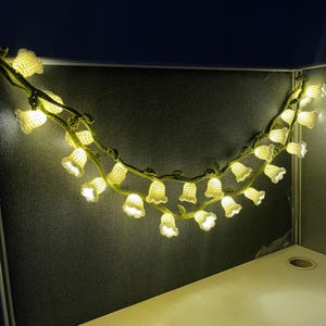 May include: A string of decorative lights featuring crocheted white bell-shaped covers attached to a green vine-like cord. The individual bell-shaped shades are illuminated from within, casting a warm glow against the dark background.