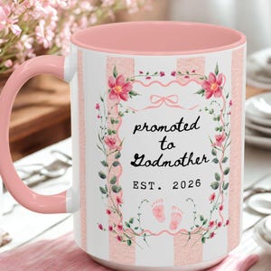 May include: A white ceramic mug with a pink interior and handle. The mug features vertical pink and white stripes and a floral wreath design with the text "promoted to Godmother EST. 2026".