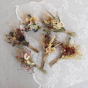 May include: Assortment of six small dried flower bouquets in various shades of brown, red, and white. The bouquets are tied with twine and arranged on a white lace tablecloth.