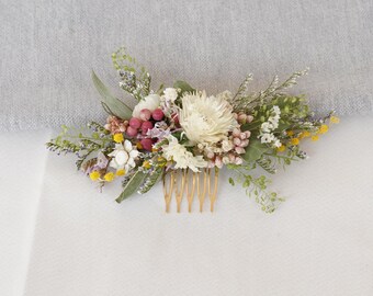 May include: A floral hair comb featuring a variety of dried flowers in shades of white, yellow, pink, and green. The flowers are arranged on a gold-colored comb, creating a delicate and natural accessory for weddings or special occasions.