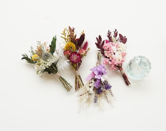 May include: Four small bouquets of dried flowers in various colors, including purple, pink, yellow, and red, tied with twine. The bouquets are arranged on a white surface.