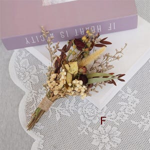 May include: A small bouquet of dried flowers tied with twine. The bouquet includes dried roses, small white flowers, and dried grasses. The bouquet is sitting on a white surface with a lace pattern.