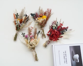 May include: Four small bouquets of dried flowers in various colors, including red, orange, yellow, and white. The bouquets are arranged on a white surface with a white book with text in the background.