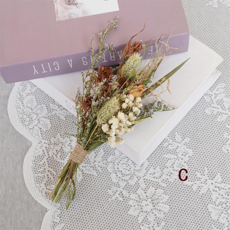 May include: A small bouquet of dried flowers in shades of brown, white, and green, tied together with twine. The bouquet is sitting on a white surface with a lace overlay.