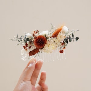 Peut inclure: Un peigne à cheveux décoré de fleurs séchées dans des tons de blanc, brun et orange. Le peigne a une base en plastique transparent et une rangée de dents.