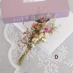May include: A small bouquet of dried flowers tied with twine. The bouquet includes pink, white, and brown flowers. The bouquet is sitting on a white lace surface.