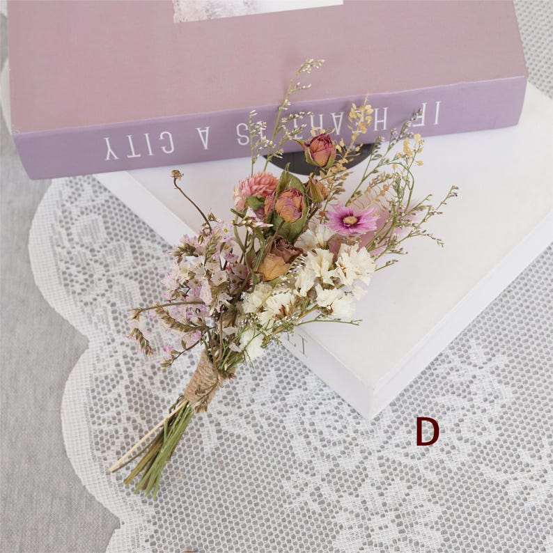 May include: A small bouquet of dried flowers tied with twine. The bouquet includes pink, white, and brown flowers. The bouquet is sitting on a white lace surface.