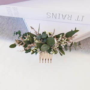 May include: A gold hair comb adorned with dried white flowers and eucalyptus leaves. The comb is a delicate accessory for a wedding or special occasion.