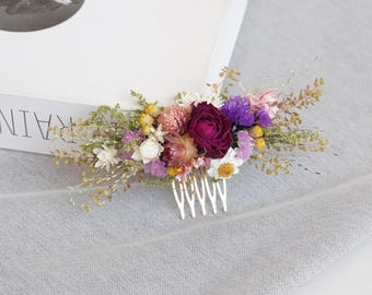 May include: A floral hair comb featuring a variety of dried flowers in shades of purple, pink, yellow, and white. The flowers are arranged on a gold-colored comb. The comb is set against a gray fabric background.