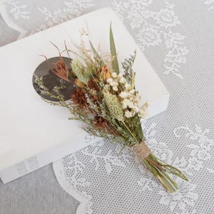 May include: A small bouquet of dried flowers in shades of brown, white, and green, tied together with twine. The bouquet is sitting on a white book with a circular image on the cover.