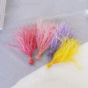May include: Four bundles of colourful, fluffy, feather-like strands. The bundles are tied together at the bottom and are arranged in a semi-circle. The colours are pink, purple, yellow, and a lighter shade of pink.