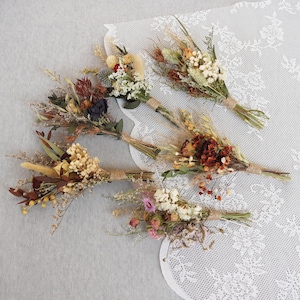 May include: Six small bouquets of dried flowers in various colors, including white, brown, orange, red, and pink. The bouquets are tied with twine and arranged on a white lace tablecloth.