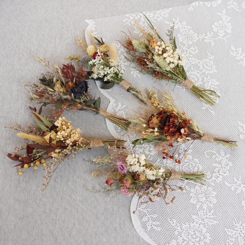 May include: Six small bouquets of dried flowers in various colors, including white, brown, orange, red, and pink. The bouquets are tied with twine and arranged on a white lace tablecloth.