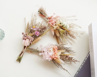 May include: Three small bouquets of dried flowers in shades of pink, white, and purple. The bouquets are tied with twine and have a rustic, bohemian aesthetic.