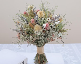 May include: A bouquet of dried flowers featuring cream-colored roses, pink globe amaranth, lavender sprigs, and various green foliage. The stems are gathered together and wrapped in a natural tan twine, creating a textured, rustic floral arrangement.