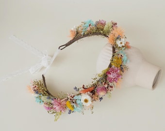 May include: A floral headband with a variety of dried flowers in shades of pink, blue, yellow, and orange. The headband is made of a brown wire and has a white lace ribbon attached.