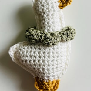 Puede incluir: Juguete de ganso blanco tejido a crochet con pico y patas amarillas y un collar verde. El ganso hecho a mano es probablemente un juguete para bebés o niños pequeños. El ganso está sobre una superficie blanca.
