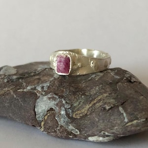 Peut inclure: Une bague en argent avec une bande texturée et une pierre précieuse rose sertie au centre.