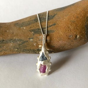 May include: A silver pendant necklace with a teardrop design. The pendant has a rough texture and incorporates a blue and a pink gemstone. The necklace is displayed on a brown stone.
