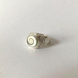 Puede incluir: Un anillo de plata con un diseño en espiral circular blanco roto y marrón. El anillo tiene una banda texturizada y un engaste liso y pulido para la espiral. Es una pieza de joyería.