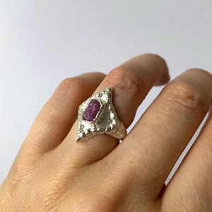 May include: A silver ring with a rough-cut, oval pink gemstone. The ring has a textured, organic design with a triangular face and a band that tapers towards the back. The gemstone is set in a bezel setting.