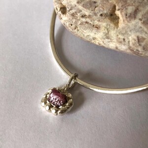 Può includere: Un braccialetto d'argento con un piccolo pendente rotondo. Il pendente presenta una pietra preziosa rosa-rossastra grezza incastonata in una cornice d'argento perlata. Il braccialetto è esposto su una superficie bianca, con una roccia di colore chiaro sullo sfondo.