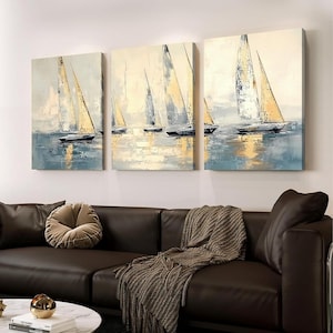 May include: Three canvas paintings depicting sailboats on the water. The artwork features a palette of blues, yellows, and whites, with the sails of the boats catching the light. The paintings are displayed above a brown leather sofa.