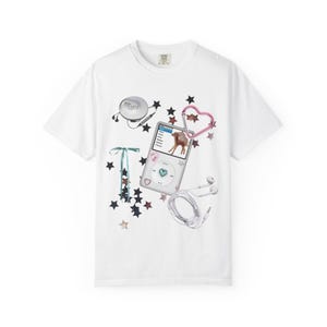 Pode incluir: Camiseta branca com um design gráfico com um leitor de MP3 vintage, fones de ouvido, um leitor de CD e um mosquetão em forma de coração rosa. O design inclui estrelas e fitas. A camiseta tem gola redonda e mangas curtas.