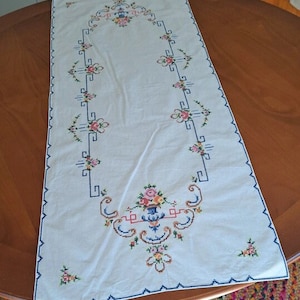 Puede incluir: Camino de mesa rectangular blanco con diseños florales y geométricos bordados en rosa, azul y amarillo. El camino de mesa tiene un borde azul y está colocado sobre una mesa de madera. El bordado presenta un jarrón de flores.