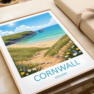 Puede incluir: Una impresión enmarcada que presenta una escena costera de Cornualles, Inglaterra. La obra de arte representa una playa de arena, agua turquesa y acantilados verdes bajo un cielo azul. La palabra "CORNWALL" está impresa en azul, con "ENGLAND" debajo.