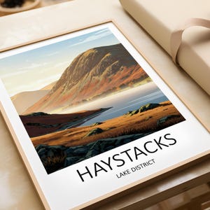 May include: Framed art print of Haystacks in the Lake District. The landscape artwork shows a mountain range with autumnal hues, a lake, and grassy areas. The print is in a light wooden frame, with the text "HAYSTACKS LAKE DISTRICT" at the bottom.